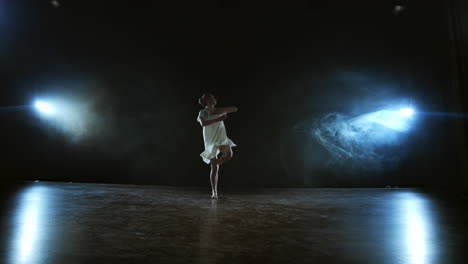a-young-girl-in-a-white-dress-dances-a-modern-ballet-makes-rotations-and-jumps-in-slow-motion-on-the-stage-with-smoke-in-spotlights-in-full-shot.-Camera-is-zooming-out-standing-behide-the-girl.
